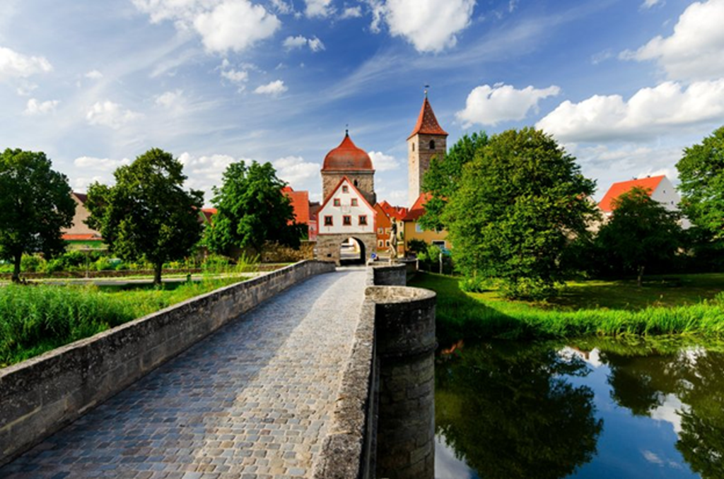 Stone bridge leading to Rothenburg’s historic gate, with towers and greenery under a partly cloudy sky.