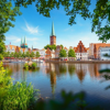 View of the historic city centre of Lübeck with colourful façades, tall church spires, and a river in the foreground, framed by green trees on a sunny day.