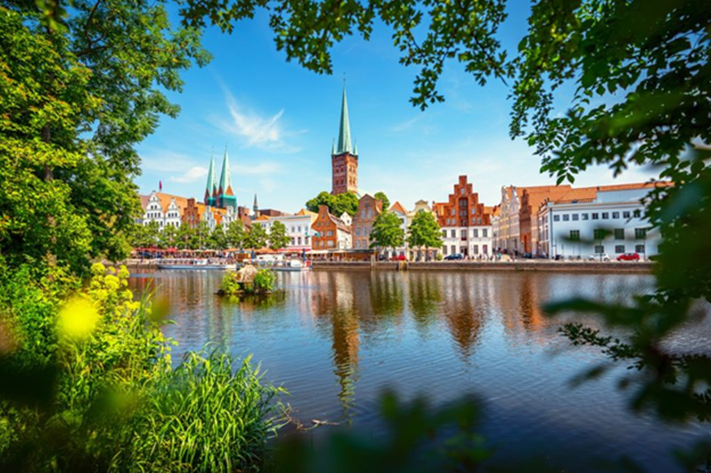View of the historic city centre of Lübeck with colourful façades, tall church spires, and a river in the foreground, framed by green trees on a sunny day.